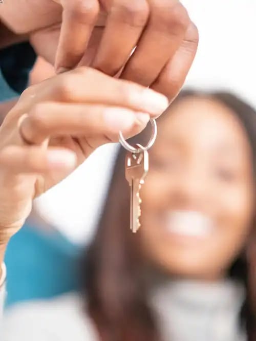 A smiling woman is blurred in the background and hands holding a house key are sharp in the foreground